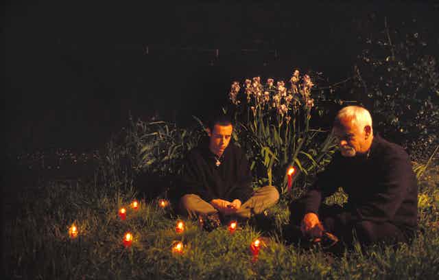 Two men, one older, sitting on grass among nature with lit candles around them.