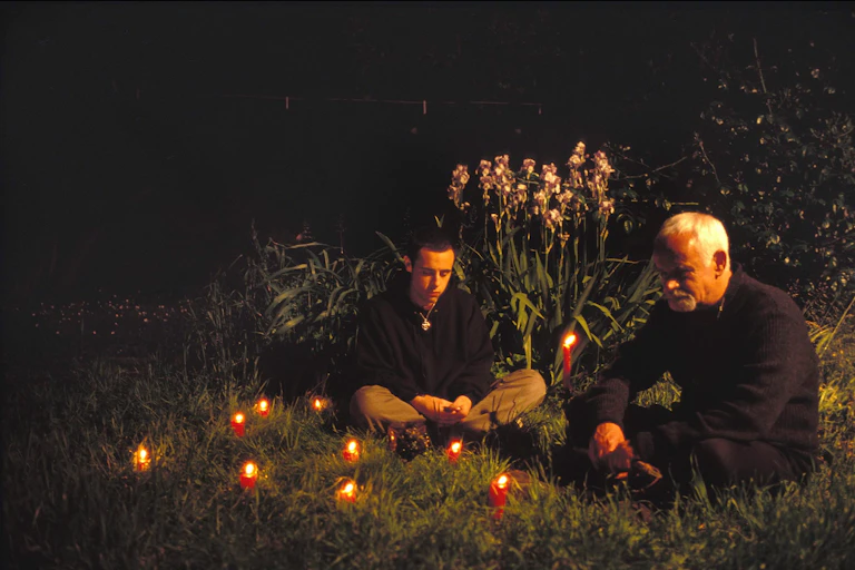 Two men, one older, sitting on grass among nature with lit candles around them.