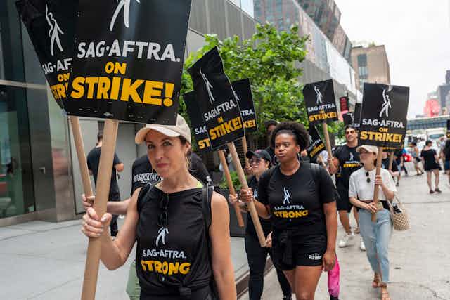 Mujeres y hombres caminando en la calle delante de un edificio y portando pancartas en las que se lee: SAG-AFTRA en huelga.