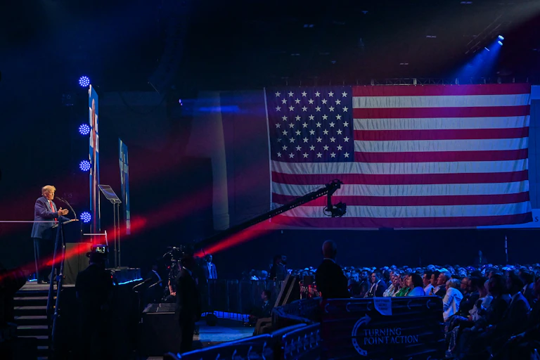 Donald Trump is seen from a distance on a stage, in front of a large American flag and a crowd of people.