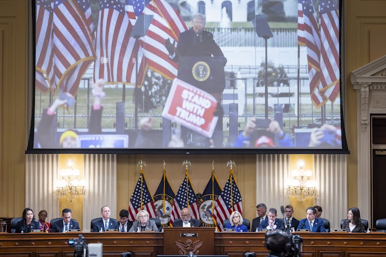 A large photo of Donald Trump is shown on a projector screen, above a row of people seated at podium with American flags behind them.