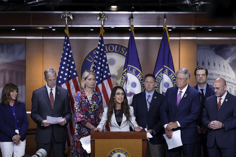 A woman with long dark hair, wearing eye glasses and a white suit speaks from a lectern. Standing behind her are suited men and women, some holding papers in their hands.