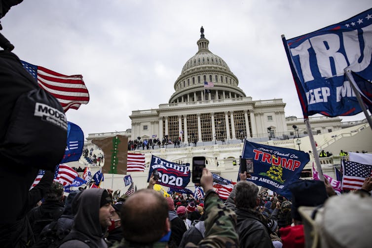 Una gran multitud de personas se paran afuera del edificio del Capitolio de los Estados Unidos y sostienen carteles que dicen Trump y banderas estadounidenses.