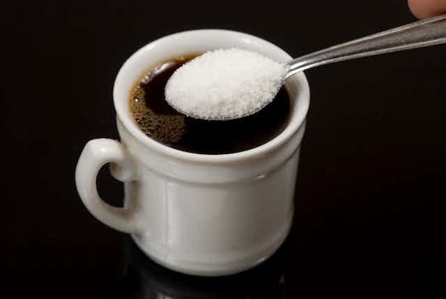 Against a black background, we see a white ceramic cup filled with coffee about to be doused with a spoonful of artificial sweetener.