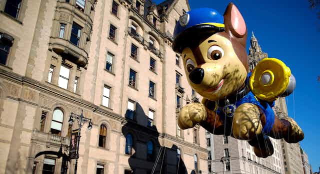 Paw Patrol dog mascot balloon seen floating above a parade against buildings and a blue sky.