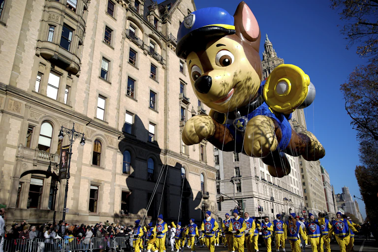 Paw Patrol dog mascot balloon seen floating above a parade against buildings and a blue sky.