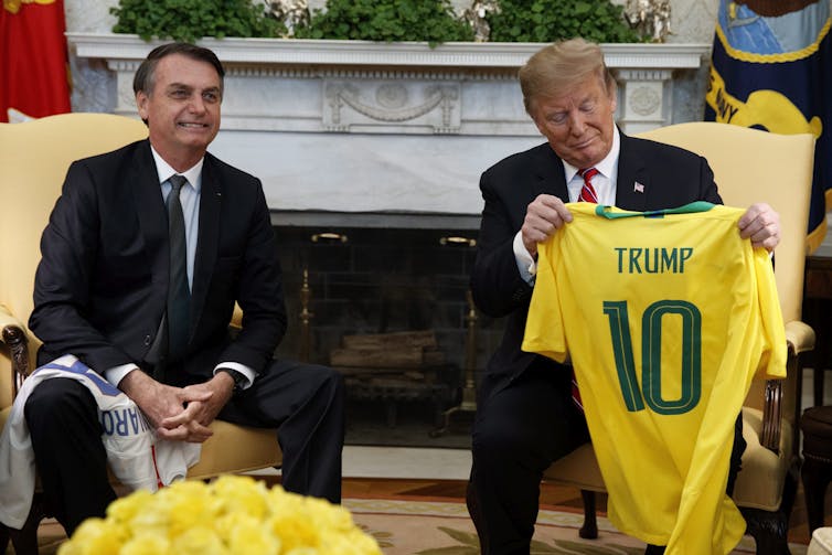 A man holds up a yellow soccer jersey with a green No. 10 while another man smiles in an armchair beside him.