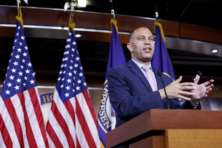 A suited man holds his hands parallel and chest-height as speaks from behind a lectern. American flags stand behind him.