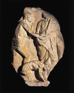 Part of a stone relief shows two people shaking hands while another crouches beneath them.