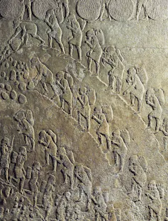 A relief shows rows of men lugging heavy items as they plod up a hill.
