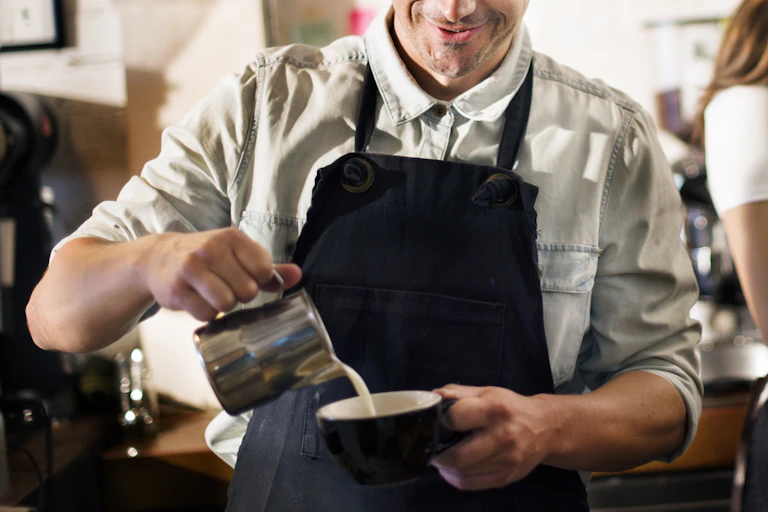 Male barista pours milk into coffee cup as female barista uses espresso machine