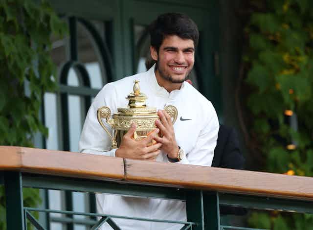 Carlos Alcaraz sonríe con un jersey blanco, sosteniendo el trofeo dorado de Wimbledon.