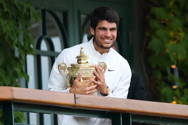 Carlos Alcaraz sonríe con un jersey blanco, sosteniendo el trofeo dorado de Wimbledon.