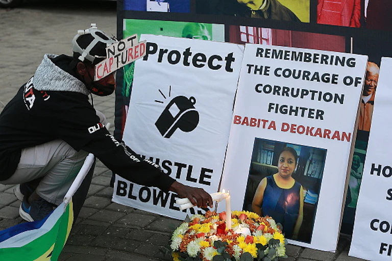 A man lights a candle in front of a poster saying 'Protect Whistleblowers' and another with the image of a woman, Babita Deokaran, a murdered whistleblower.