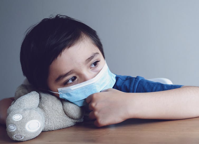 Child wearing medical face mask lying his head on teddy bear.