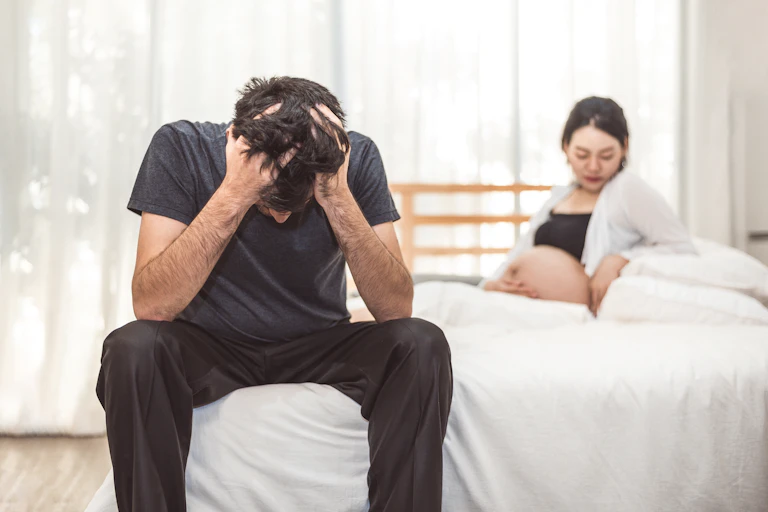 A man sits on the edge of a bed with his head in his hands. A pregnant woman is in the bed.