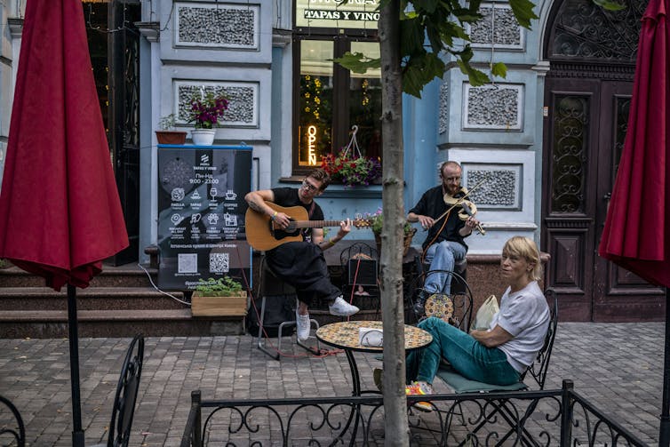 Dos hombres tocan una guitarra y un violín en una calle aparentemente vacía, con una mujer sentada en una silla cerca.
