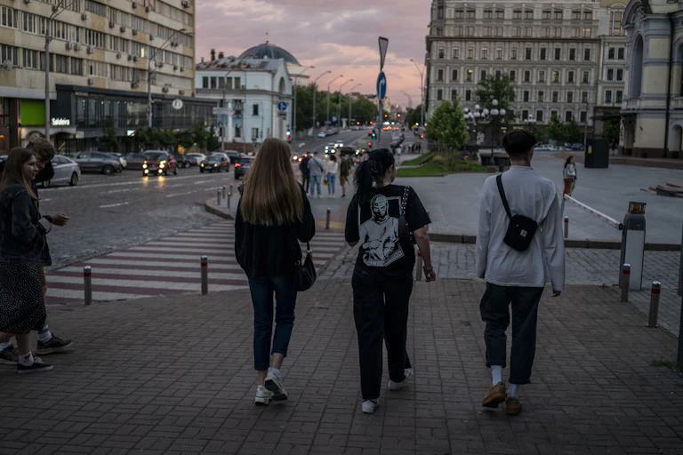 The backs of people walking across a street are seen, as the sun appears to set near buildings in the distance.
