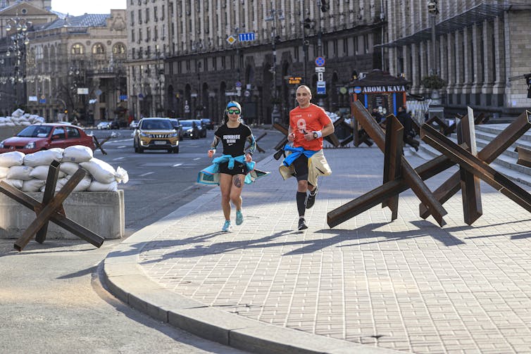 People are seen running in athletic clothing past large sandbags and a teal cross.