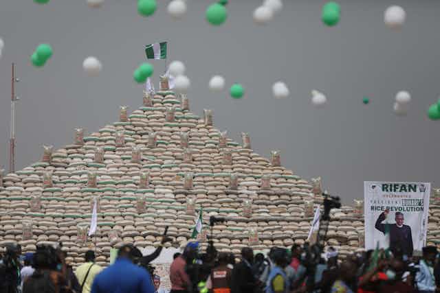 People gather to look at some bags of rice stacked in a pyramid.