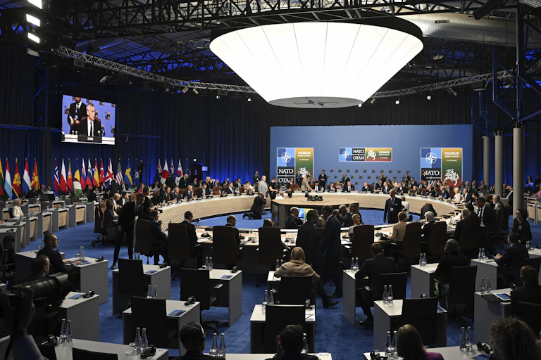 A large room with a circular table in the middle with people at seats around it, and at desks on the outside of the table.