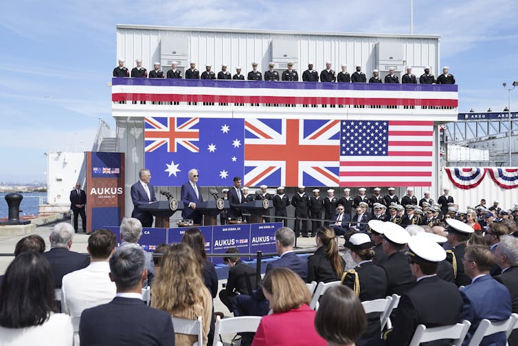 Una multitud mira un gran cartel con las banderas de Australia, Reino Unido y Estados Unidos, mientras tres hombres se paran en los atriles justo debajo.