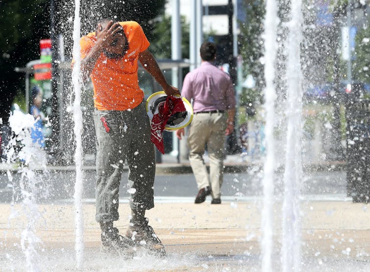 A construction worker cools his head in the stream of a fountain outside an office building.