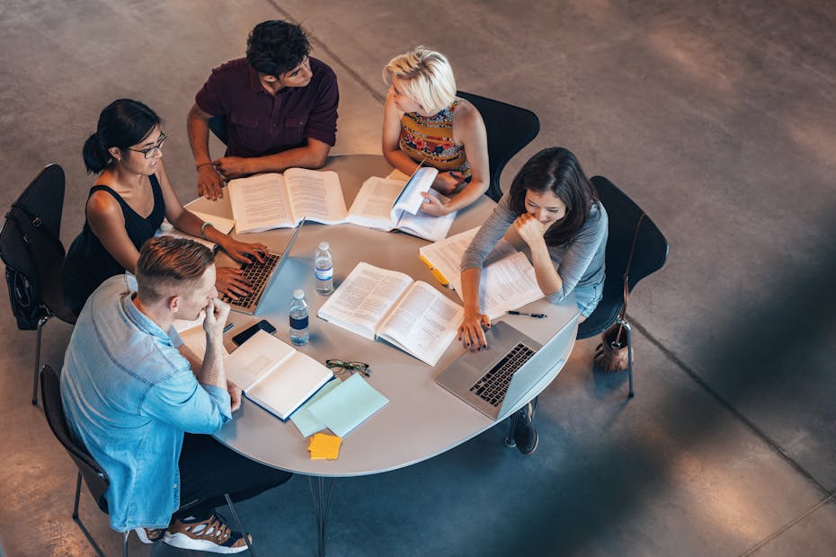 A group of people sit at a table with books and laptops.