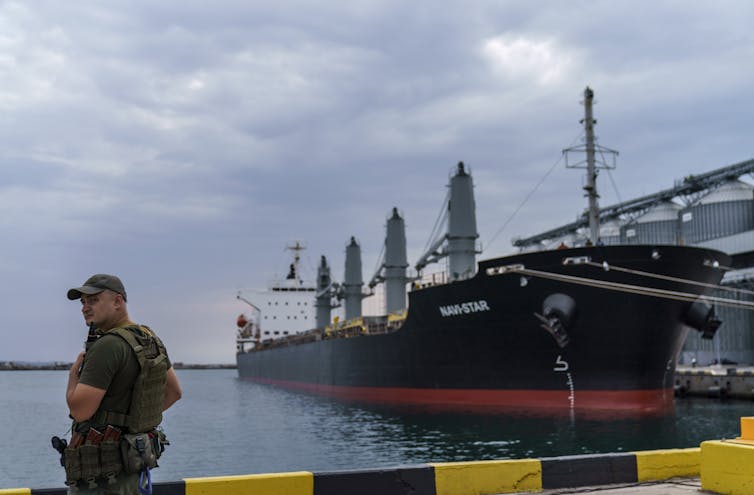 a man in military gear stands near the water and a big black ship docked