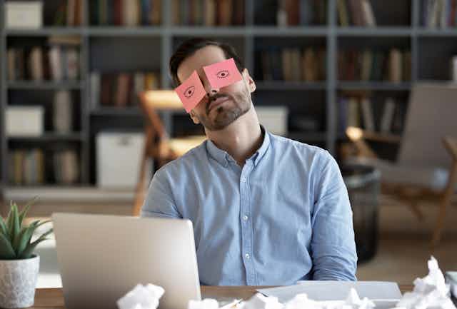 Un hombre frente a un ordenador con dos post-its con un dibujo de unos ojos abiertos cubriéndole sus propios ojos.