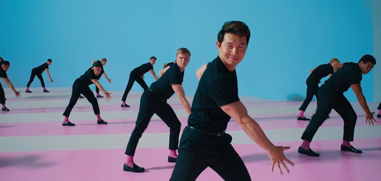 Simu Liu wearing all black along with many other Kens in a dance sequence.