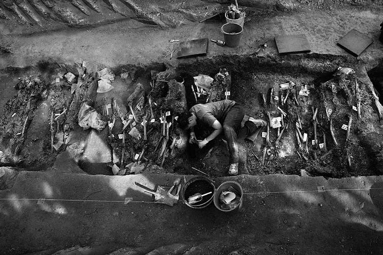 Fotografía de una fosa común con huesos marcados con señales y con un trabajador que sigue excavando.