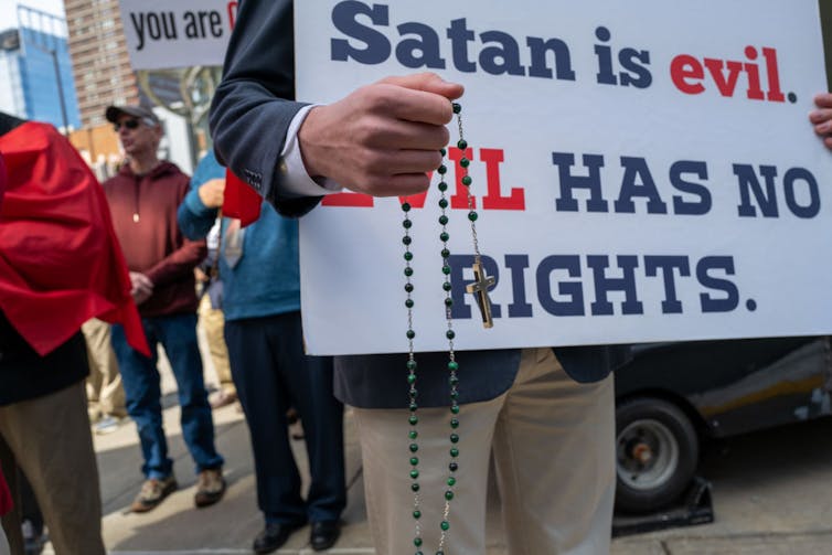 A handful of people stand at a protest, with one holding a rosary and a sign that says, 'Satan is evil. EVIL HAS NO RIGHTS.'