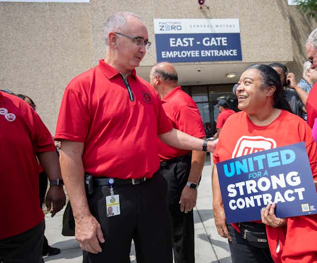 A tall man in a red shirt greets a shorter woman holding a sign that says 'United for a Strong Contract.'