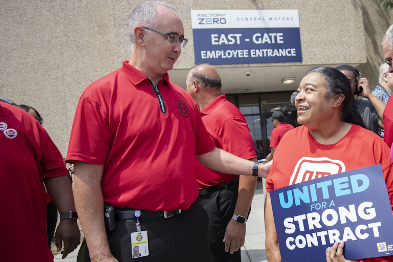 A tall man in a red shirt greets a shorter woman holding a sign that says 'United for a Strong Contract.'