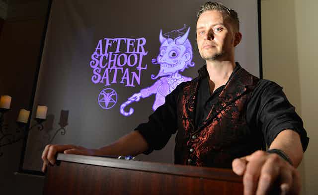 A blond man in a black shirt and a shiny red vest stands at a podium in front of a sign that says, in purple font, 'After School Satan.'
