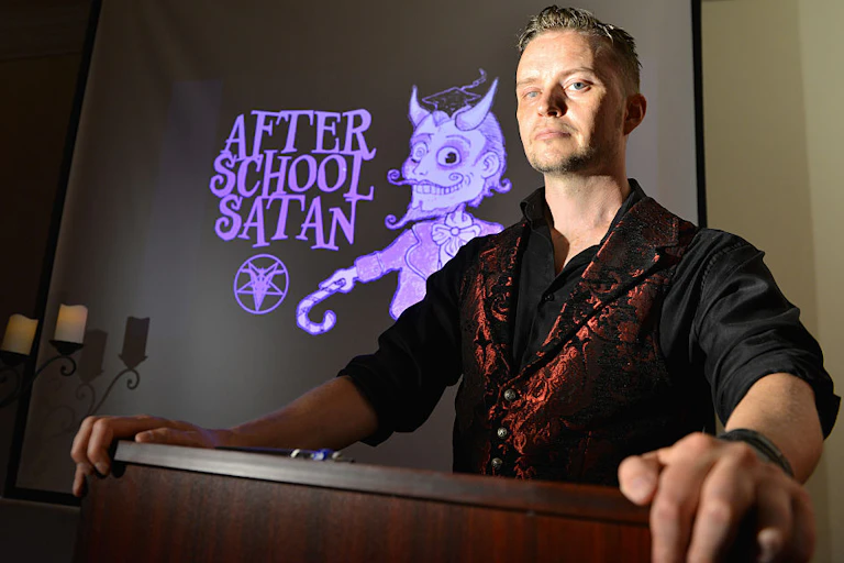 A blond man in a black shirt and a shiny red vest stands at a podium in front of a sign that says, in purple font, 'After School Satan.'