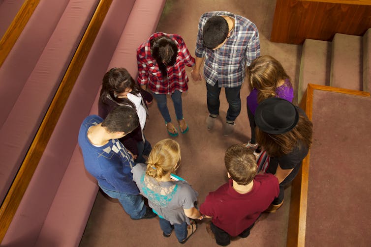 Eight teenagers, seen from above,   Istand in an empty church while holding hands and bowing their heads.