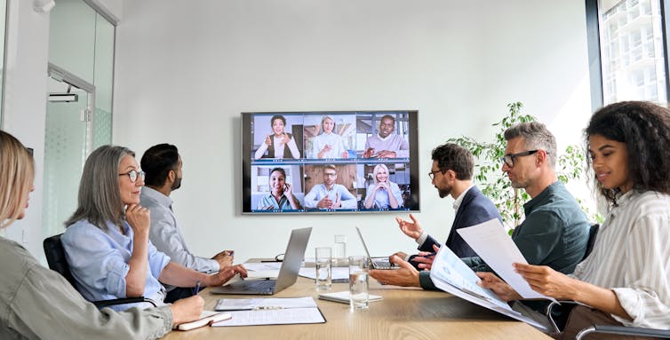 Group of people in work clothes around a table, looking at papers, with a screen showing a video call with other people in the background.