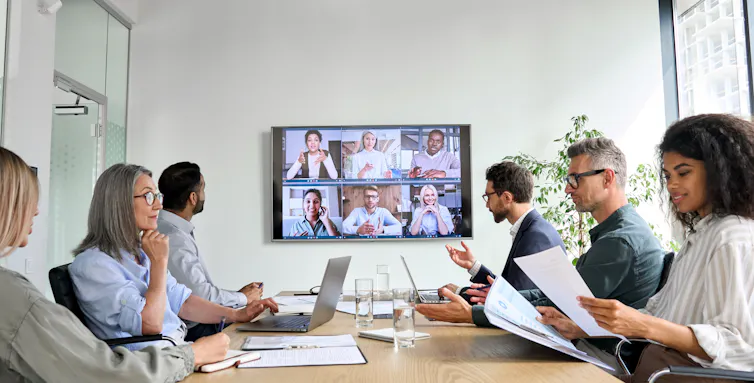 Group of people in work clothes around a table, looking at papers, with a screen showing a video call with other people in the background.