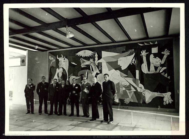 Varios hombres trajeados posan delante del inmenso mural 'Guernica' creado por Pablo Picasso, en el que, con estilo cubista, plasma el bombardeo de la ciudad de Guernica durante la guerra civil.