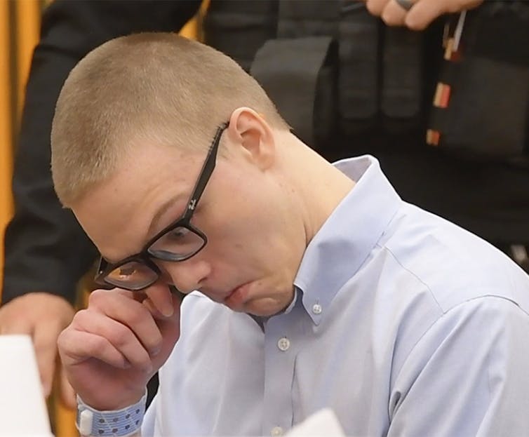 A young man with buzz-cut hair and black glasses wipes his eye, while wearing a button-up shirt.