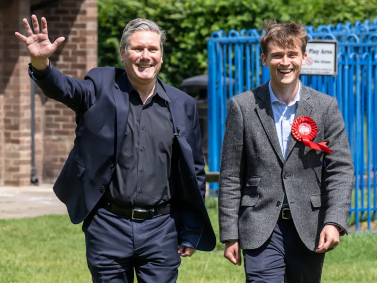 Keir Starmer smiles and waves, walking next to Keir Mather, smiling and wearing a Labour ribbon on his lapel.