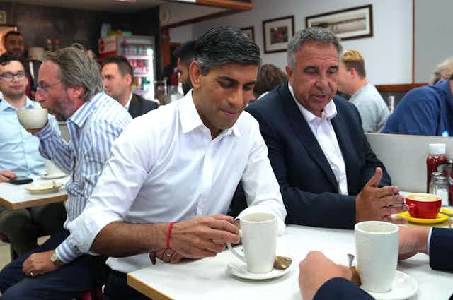 Rishi Sunak and Steve Tuckwell sitting side by side in a busy cafe, Sunak about to lift up a cup of tea
