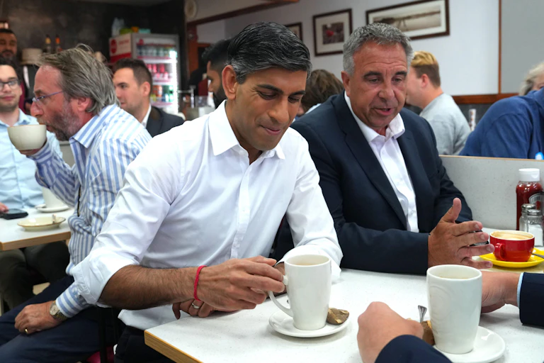 Rishi Sunak and Steve Tuckwell sitting side by side in a busy cafe, Sunak about to lift up a cup of tea
