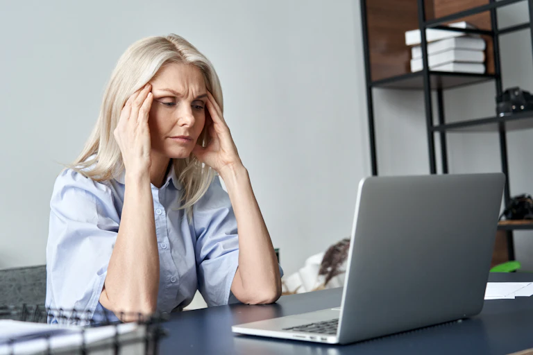 Woman with hands to head, looking distressed, in front of laptop.