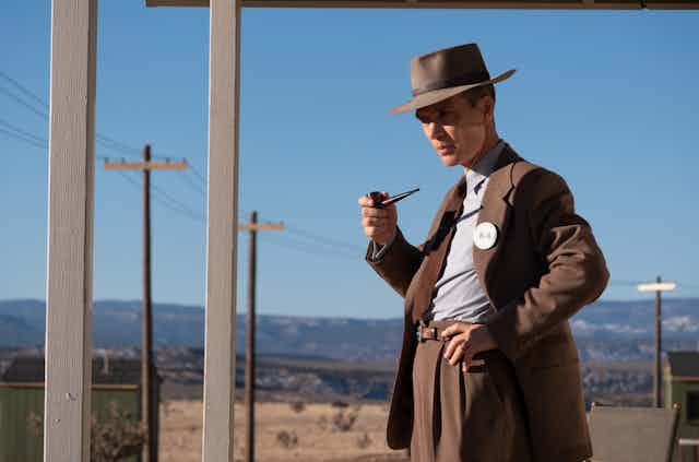 Cillian Murphy  smoking a pipe wearing a brown suit and matching hat, with telephone poles in the background.