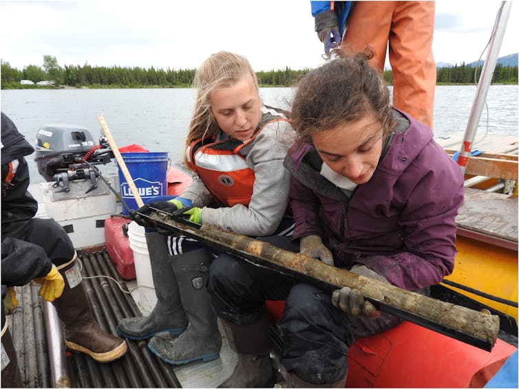 Two female scientists aboard a boat examine a sediment core, with the layers clearly visible.