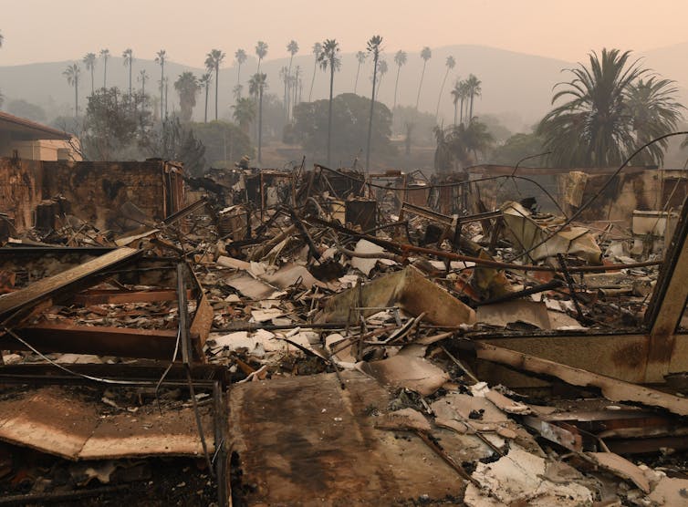 Palm trees stand above the wreckage of a fire-burned building and homes. The air is still smoky.