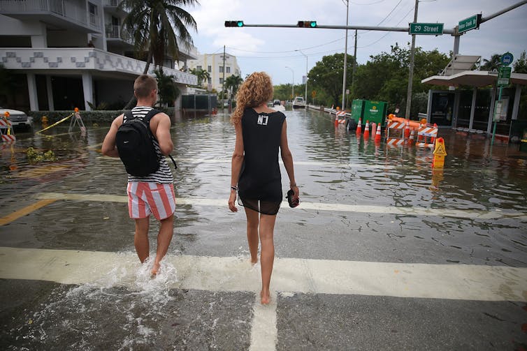 A woman in a dress carrying her shoes and a man in red and white striped shorts walk down a street that is filled with water to above their ankles.